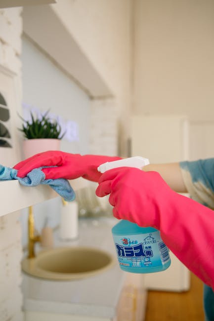 A woman in an orange apron is engaged in surface cleaning in a modern kitchen, using a blue cloth or spray bottle. The kitchen features a sleek, white marble backsplash and countertops with subtle gre