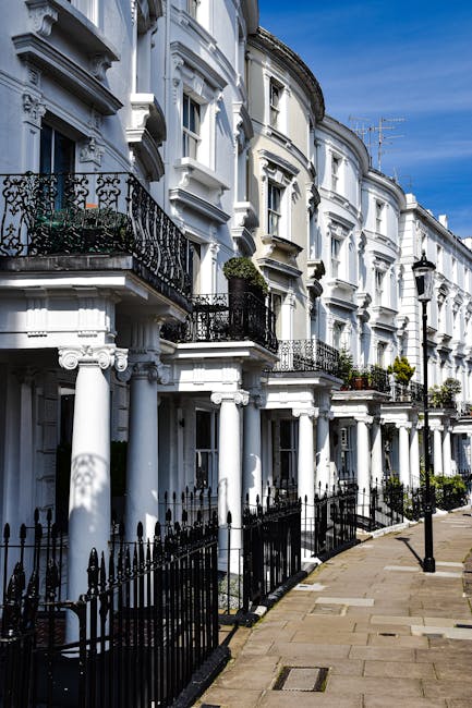 A row of white Victorian-style terraced houses with ornate cornices and decorative moldings, featuring small black wrought iron balconies on the first floor and large sash windows. The buildings are set along a paved sidewalk with a black iron fence in front, and have lush green plants and flowers on some balconies. Bright daylight illuminates the scene, highlighting the clean, well-maintained facades and the clear blue sky above. Pimlico Cleaners specializes in surface cleaning and deep cleaning services for residential properties, ensuring thorough sanitisation and hygiene in homes and flats, including those in the SW1V area. This image exemplifies the importance of regular domestic cleaning to maintain a fresh, dust-free environment in period terraced housing.