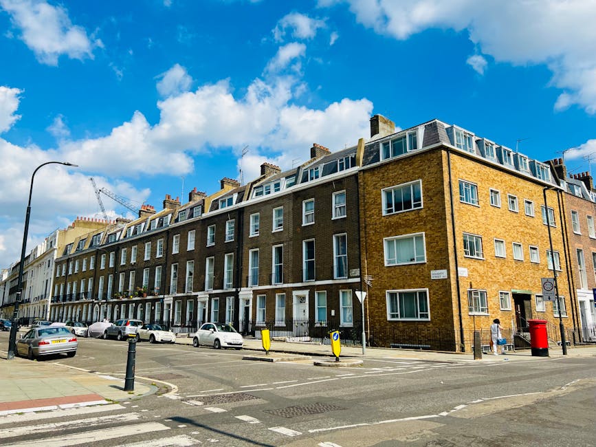 Photograph of a row of three to four-story terraced residential buildings in Pimlico, London, constructed with yellow and brown bricks, featuring uniform white-framed windows and flat roofs with dormer windows. The street in front shows parked cars, a pedestrian crossing, and a red post box near the corner. Bright daylight highlights the clean and well-maintained exterior surfaces, with a clear blue sky and scattered white clouds overhead. Pimlico Cleaners provides professional surface cleaning and deep cleaning services to maintain the hygiene and presentation of such properties, as referenced in the SW1V end-of-tenancy cleaning checklist for Pimlico flats, PIMLICO.