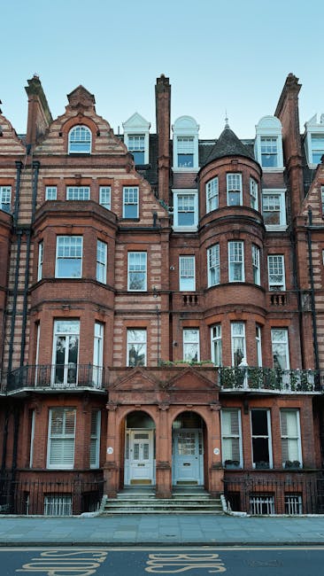 A row of white Victorian-style terraced houses with ornate cornices and decorative moldings, featuring small black wrought iron balconies on the first floor and large sash windows. The buildings are set along a paved sidewalk with a black iron fence in front, and have lush green plants and flowers on some balconies. Bright daylight illuminates the scene, highlighting the clean, well-maintained facades and the clear blue sky above. Pimlico Cleaners specializes in surface cleaning and deep cleaning services for residential properties, ensuring thorough sanitisation and hygiene in homes and flats, including those in the SW1V area. This image exemplifies the importance of regular domestic cleaning to maintain a fresh, dust-free environment in period terraced housing.